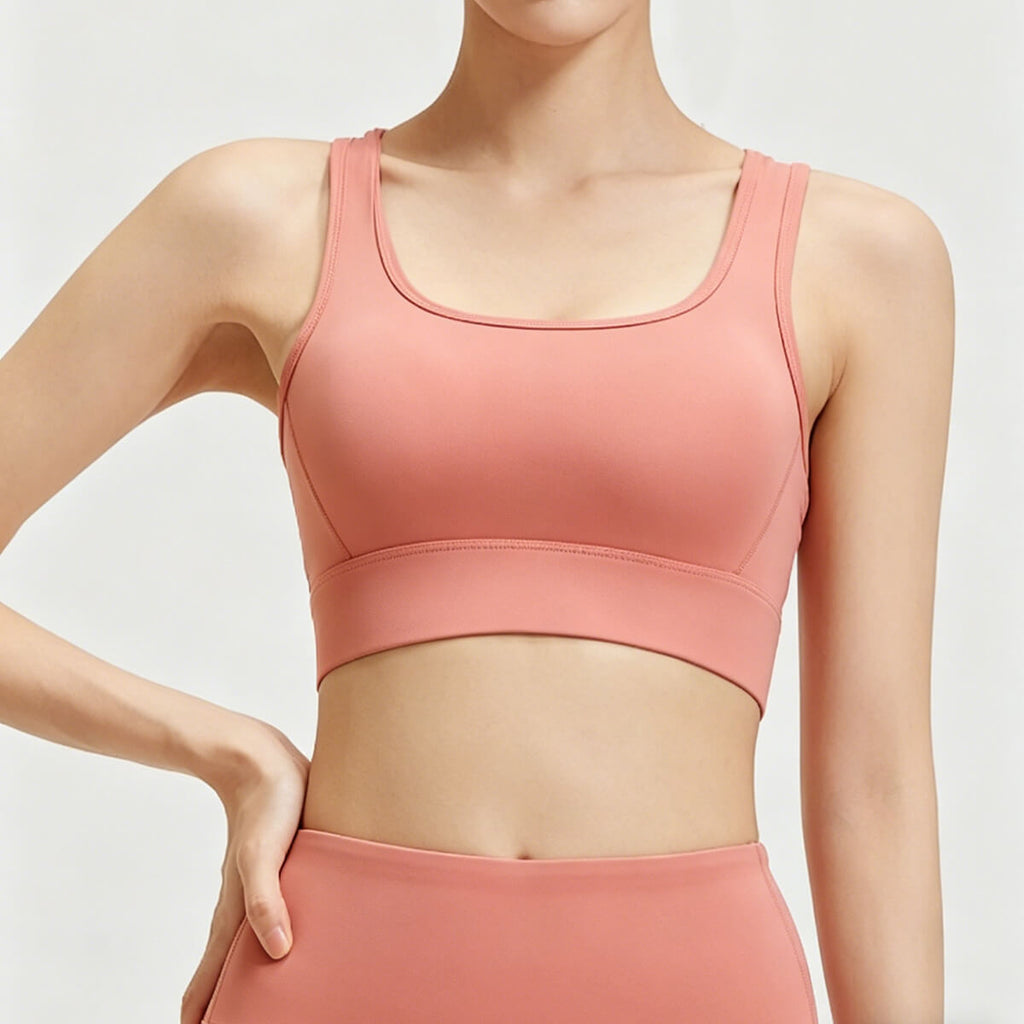 Front view of a women's coral pink scoop neck sports bra featuring wide supportive straps and a seamless finish for yoga and fitness