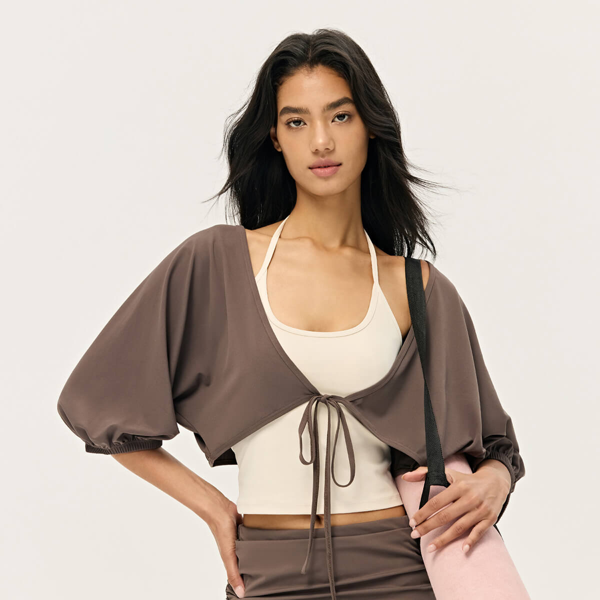 A model in an espresso brown cropped yoga shrug and matching sports bra, carrying a pink yoga mat.