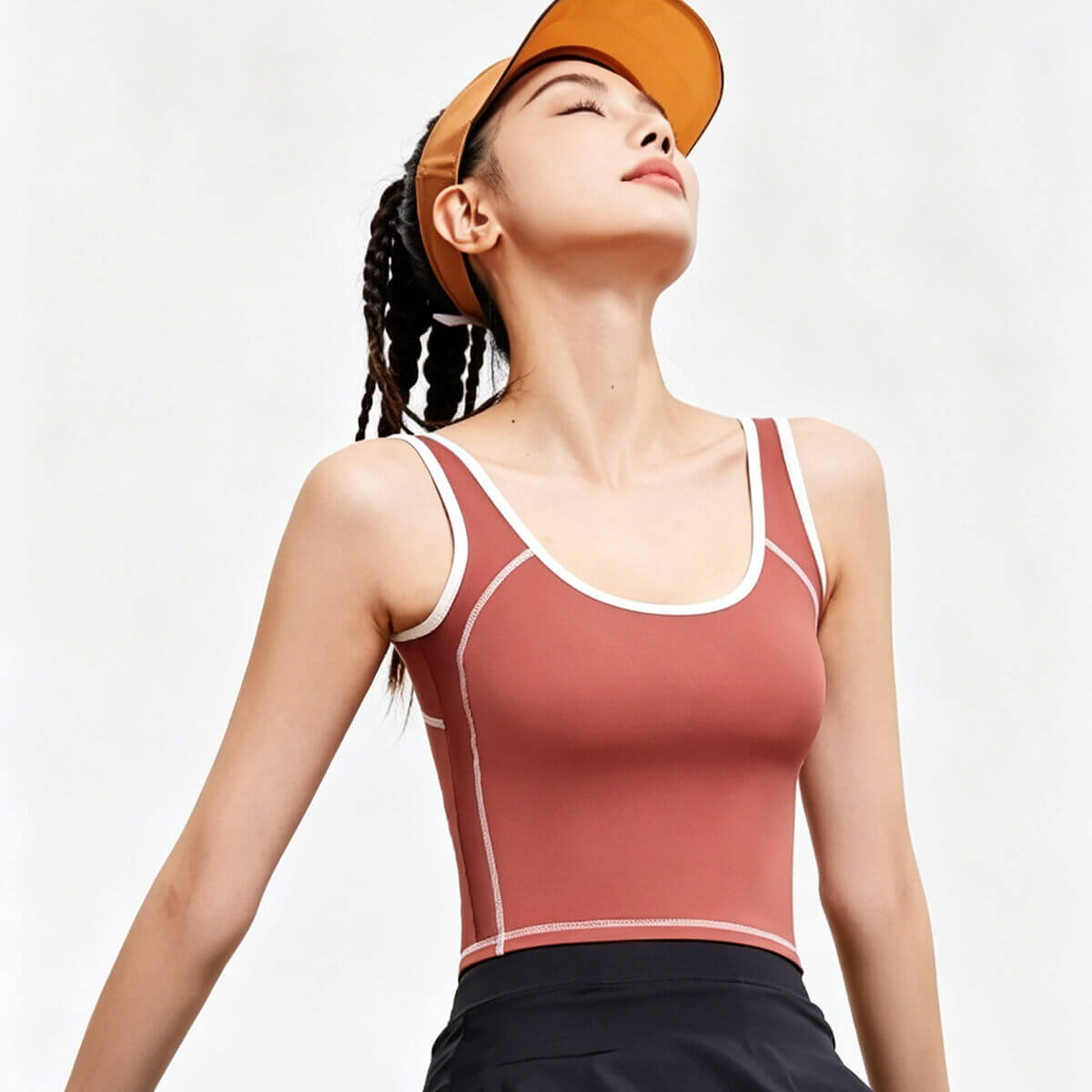 A woman wearing a rust red yoga crop top with white contrast piping and an orange sun visor, designed for outdoor tennis or running