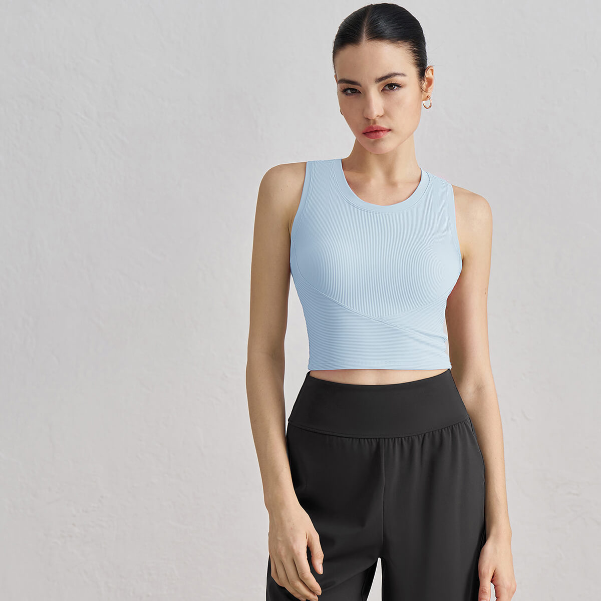 Front view of a woman wearing a sky blue ribbed high-neck cropped tank top with a flattering criss-cross waist detail.