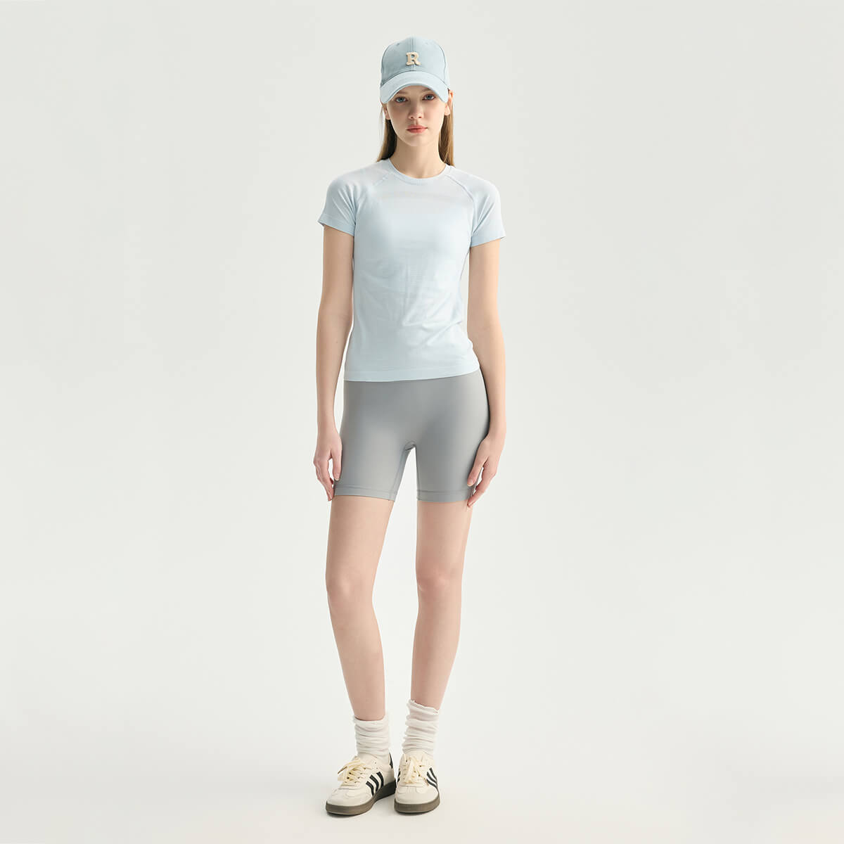 Full body view of a model in an ice blue athletic tee and grey biker shorts with sneakers and socks.