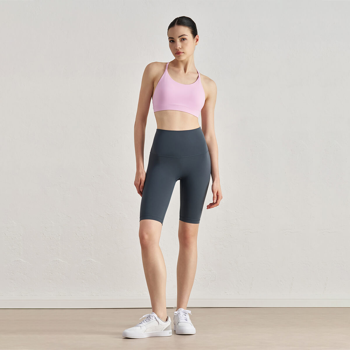 Full-body view of a model in deep grey athletic shorts and a light pink sports bra with sneakers