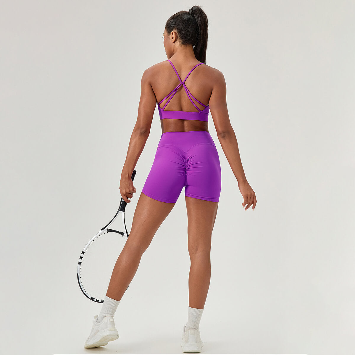 Back view of a matching purple workout set with a strappy bra and high-waisted scrunch butt shorts, holding a tennis racket.