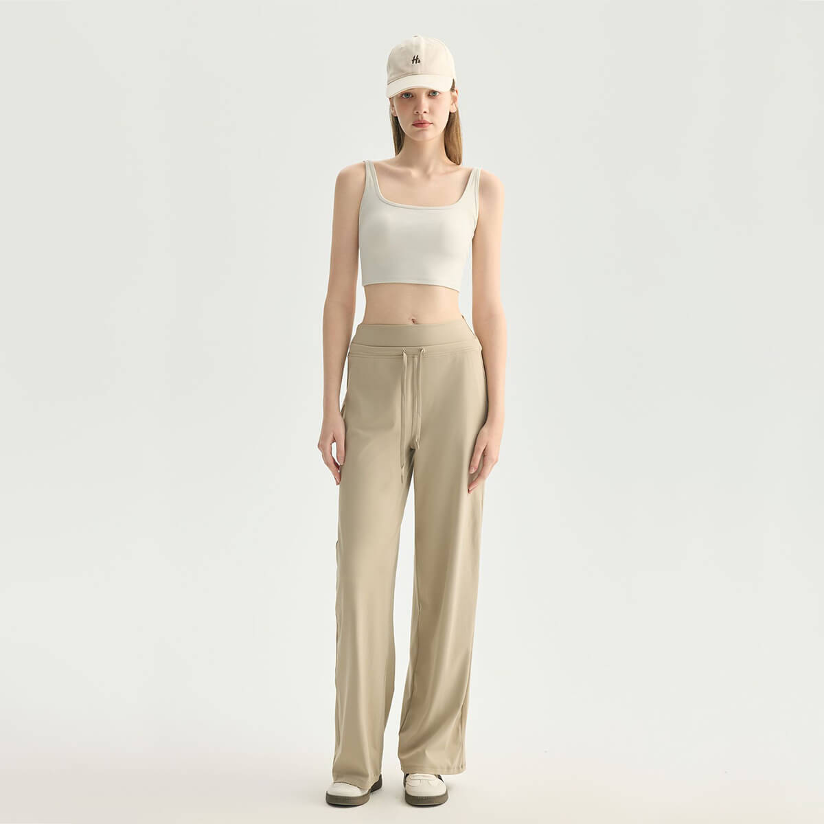 Full body front view of a woman wearing a cream square neck crop tank top and khaki wide-leg yoga pants with a baseball cap for an athleisure look.