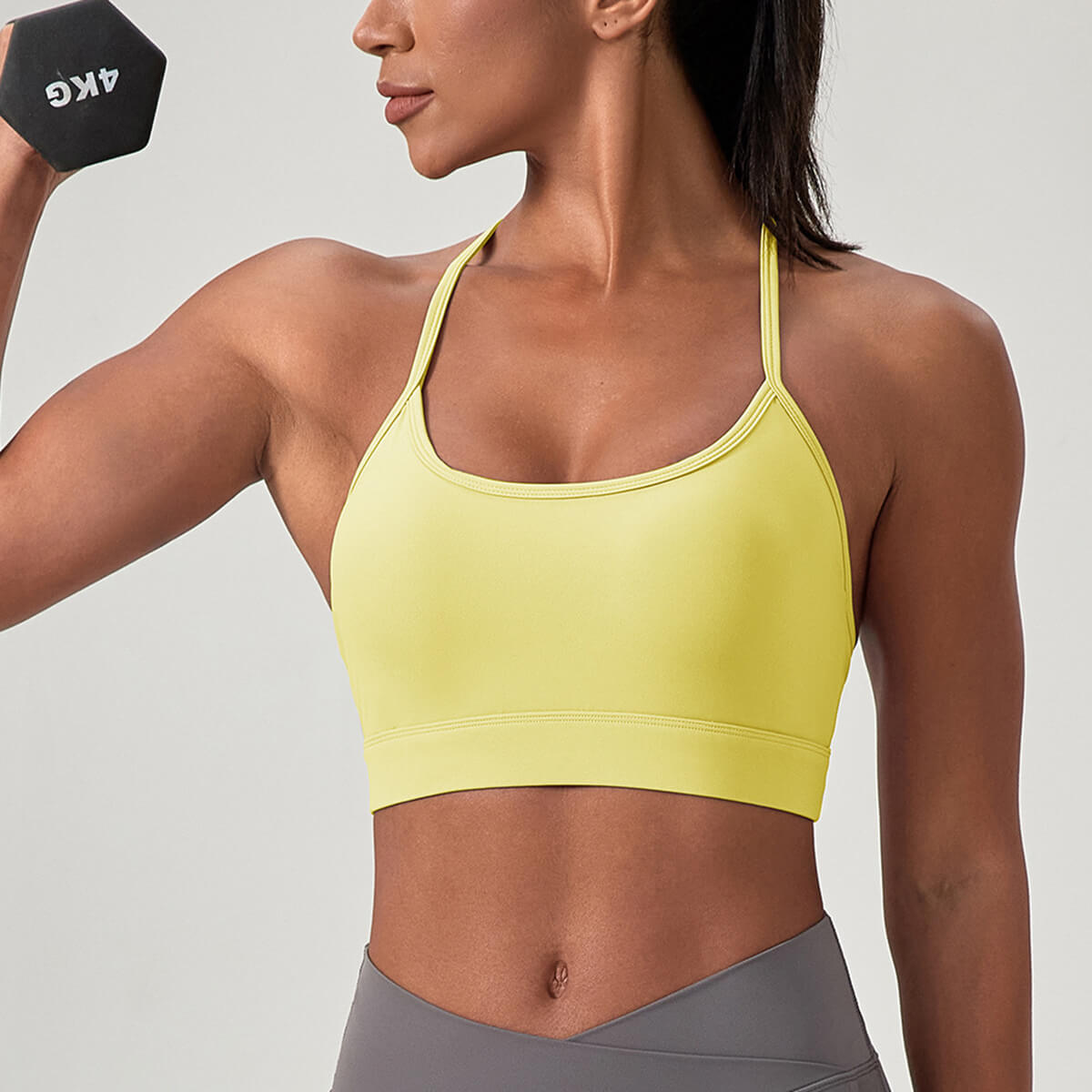 A woman holding a dumbbell wearing a pale yellow padded sports bra, demonstrating support for strength training and bodybuilding.